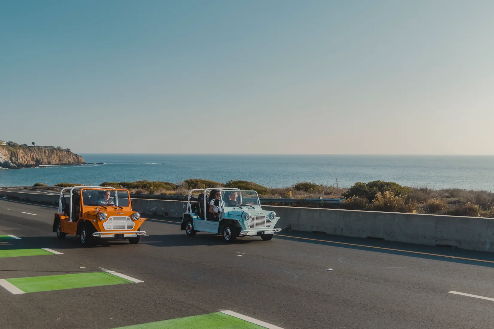 Two Moke EVs driving along a coastal highway with ocean cliffs in the background